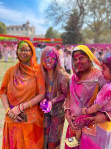 Three women and a child, covered in vibrant Holi colors, smile joyfully outdoors. They wear traditional attire, surrounded by others celebrating Holi.