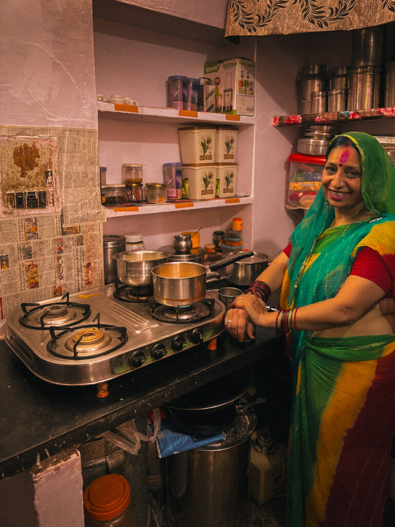 A woman in a colorful sari stands smiling in a small kitchen. The room is cozy, with a stovetop, pots, and shelves filled with jars and tins, conveying a warm, welcoming atmosphere.