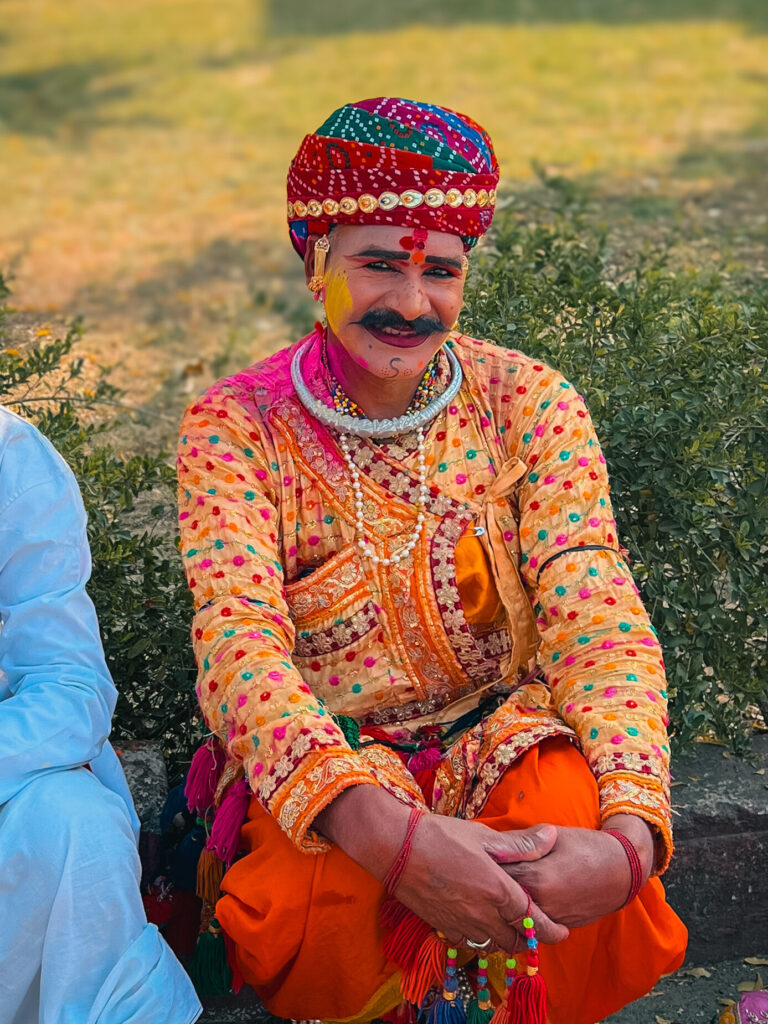 A person in vibrant traditional attire with a colorful turban and painted mustache smiles, sitting outdoors on a sunny day, conveying joy and festivity.