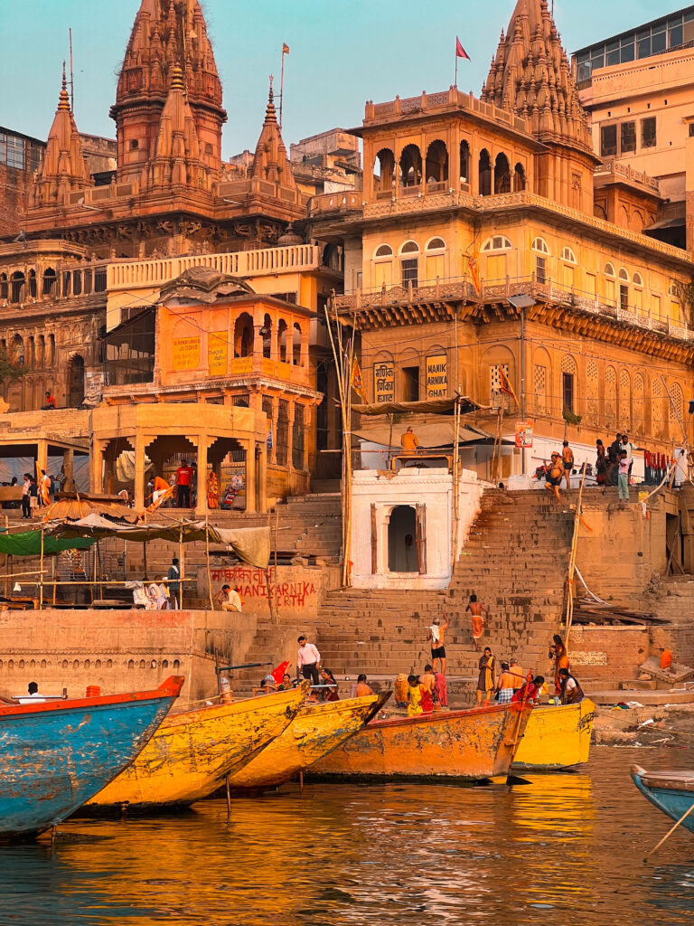 A vibrant riverside scene in Varanasi at sunset, with ornate temple buildings, steps leading to the water, and colorful boats along the Ganges River.