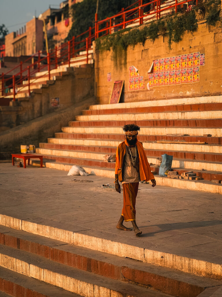 A person wearing an orange outfit and turban walks down sunlit stone steps beside a wall with vibrant posters. The scene conveys a peaceful, warm atmosphere.
