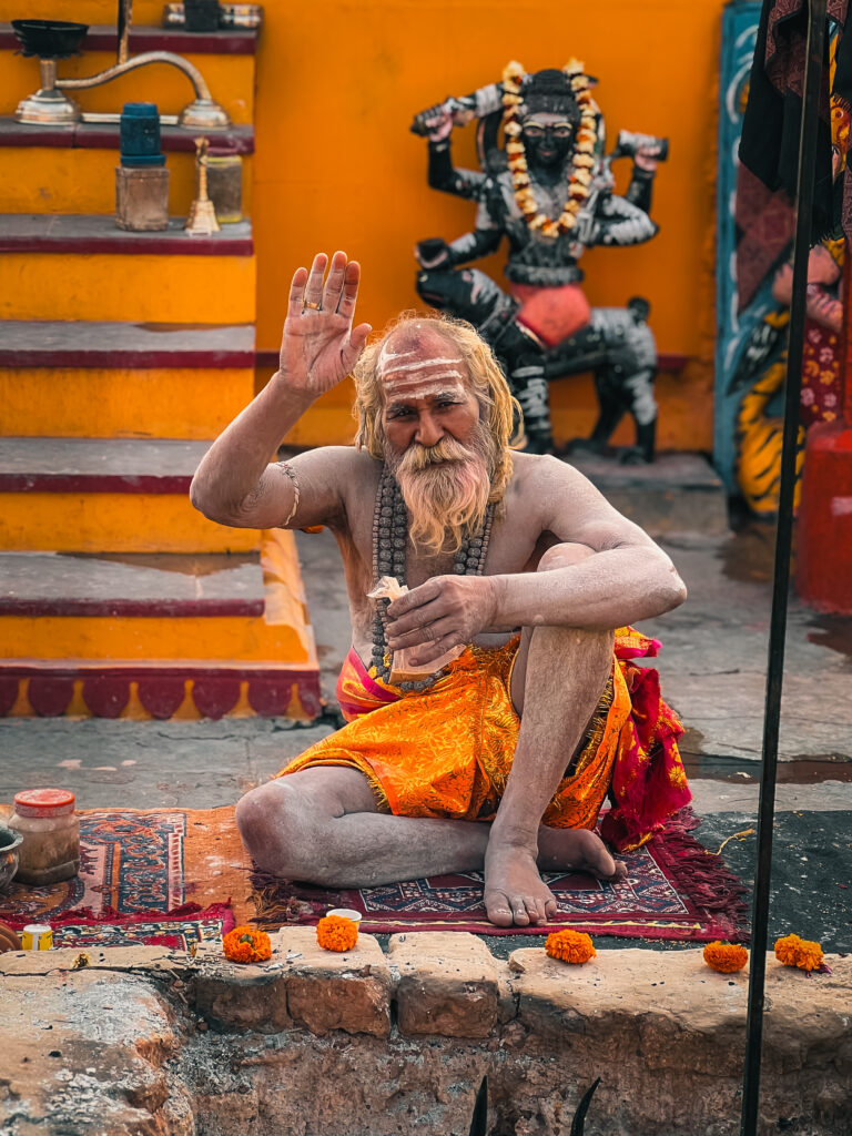 Seated on a vibrant rug, an elderly man with long gray hair waves, wearing orange robes. Behind him, colorful statues and offerings create a spiritual atmosphere.