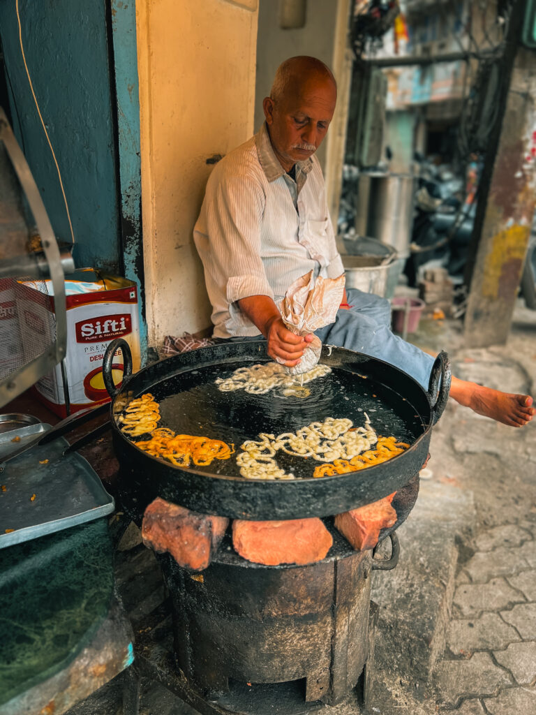Elderly man making jalebi at a street stall, using a large black pan on rustic stove. Surroundings are vibrant and colorful with a relaxed atmosphere.
