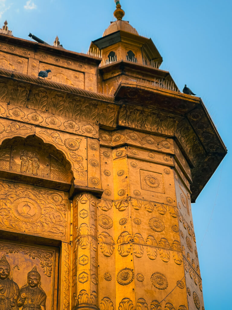 Ornate golden temple facade adorned with intricate carvings against a clear blue sky. Birds perch on the roof, adding a serene, timeless feel to the scene.