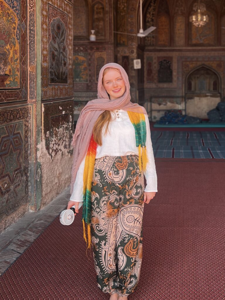 A woman in colorful attire stands in a richly decorated historic building, smiling. The atmosphere is warm with intricate patterns on the walls.