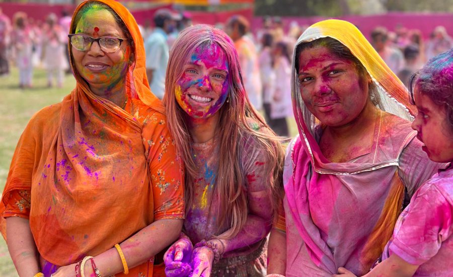 Three women and a child, covered in vibrant Holi colors, smile joyfully outdoors. They wear traditional attire, surrounded by others celebrating Holi.